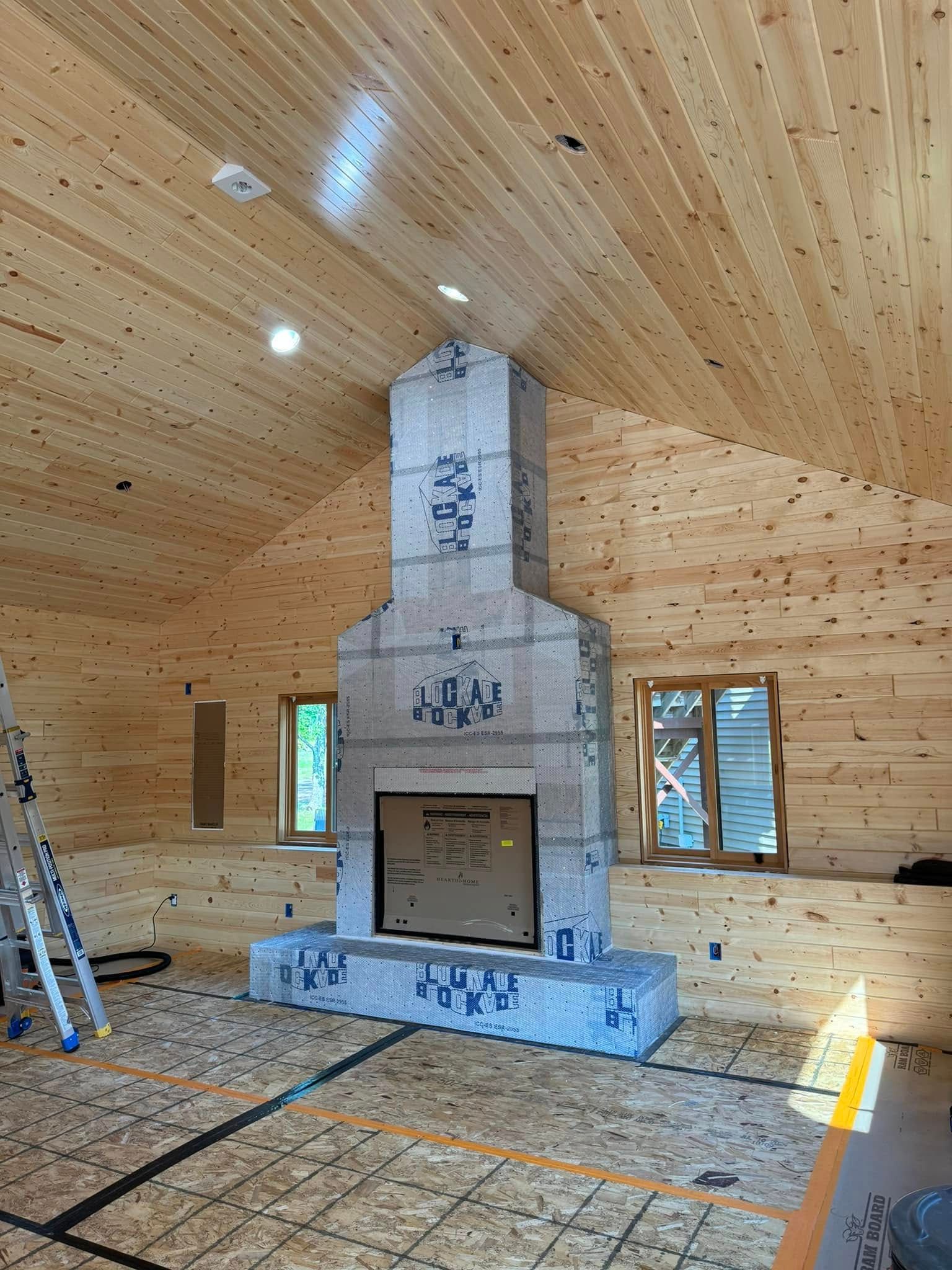 Unfinished interior: Stone fireplace under construction in a wood-paneled room with windows and sloped ceiling.