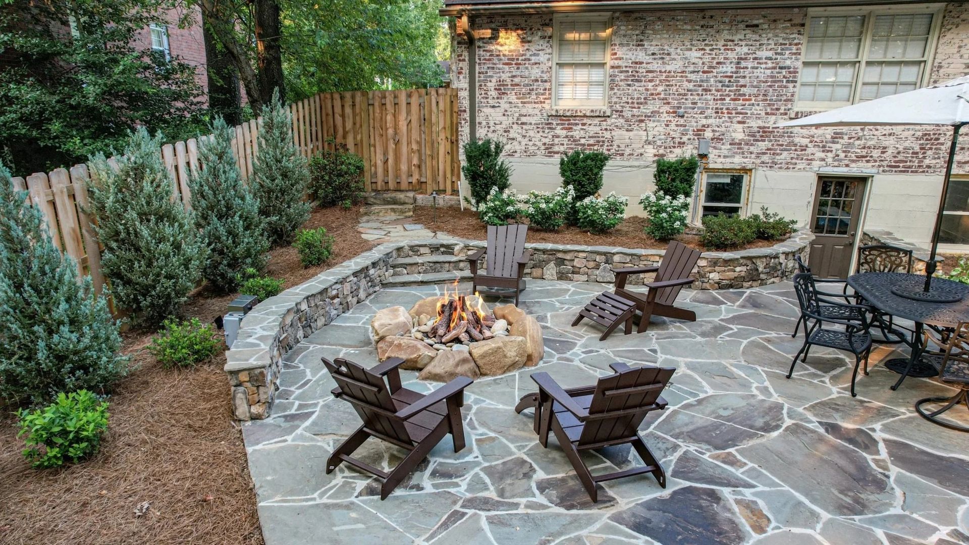 Backyard patio with fire pit, stone patio, wooden chairs, and landscaping.