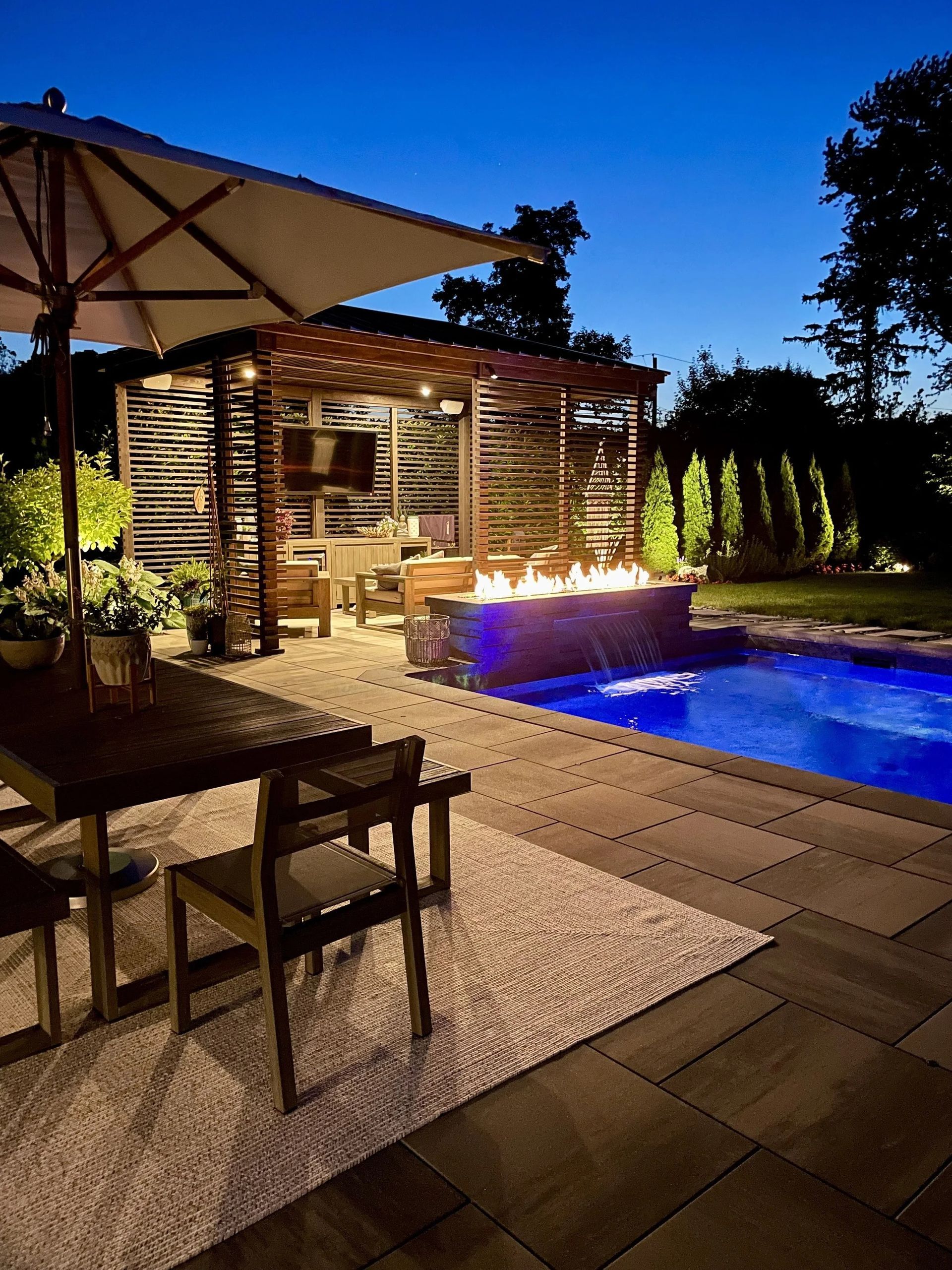 Outdoor evening scene: illuminated pool, pergola with fireplace and TV, dining table under umbrella.