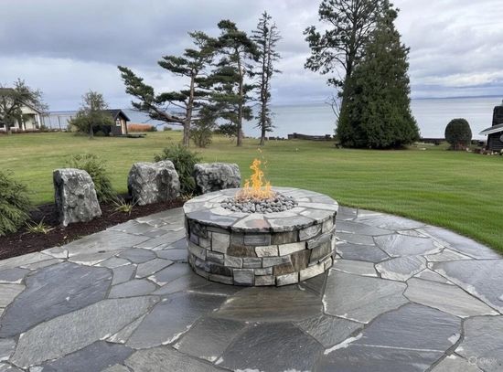 Fire pit with flames on a stone patio overlooking a lake and grassy lawn.