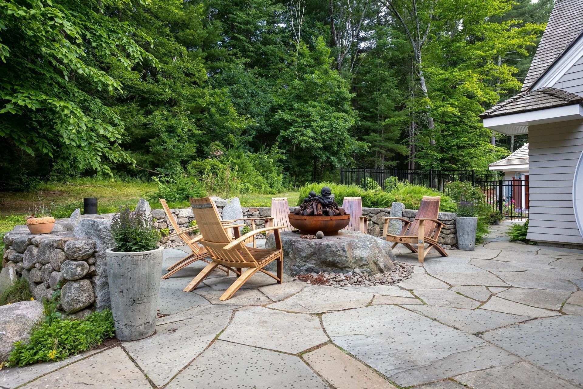 Stone patio with seating area, fire pit, and stone wall backdrop. Surrounded by trees.