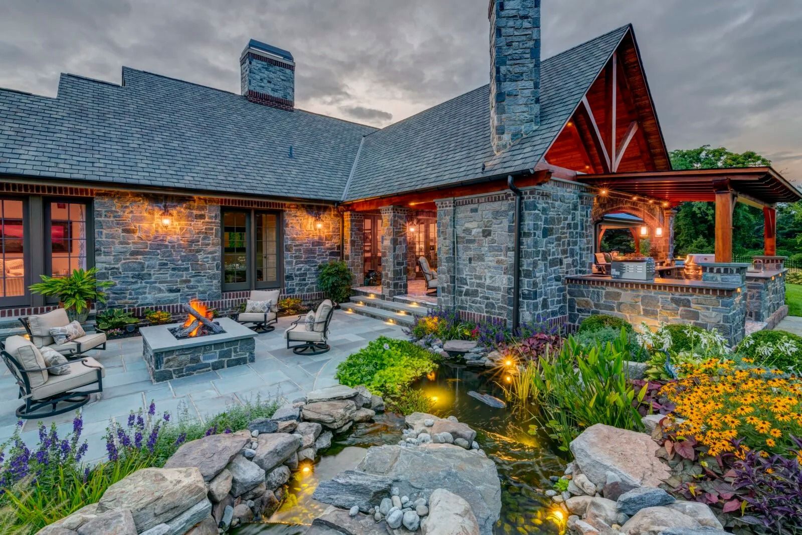 Stone house exterior with patio, fire pit, pond, and outdoor kitchen, illuminated at dusk.