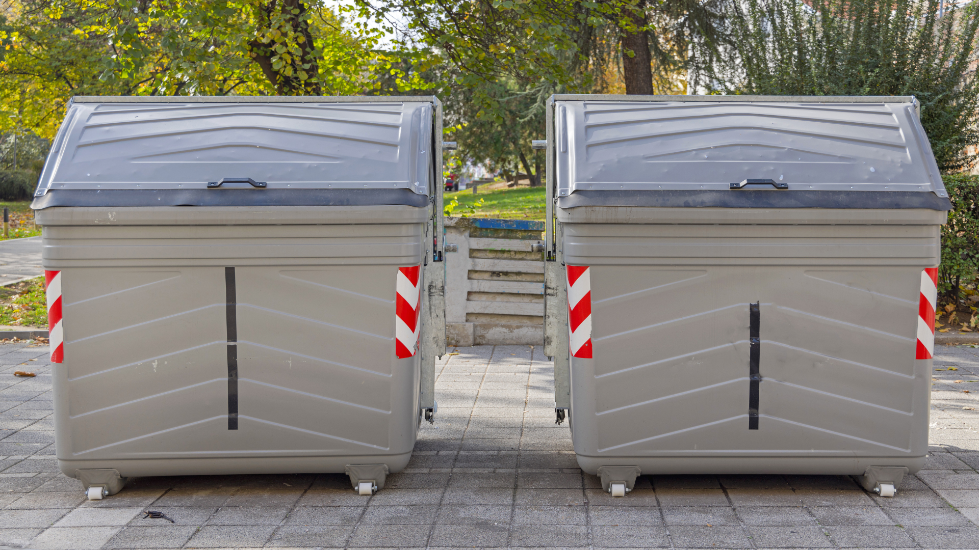 Two large, gray trash containers side-by-side outdoors with red and white striped markers.