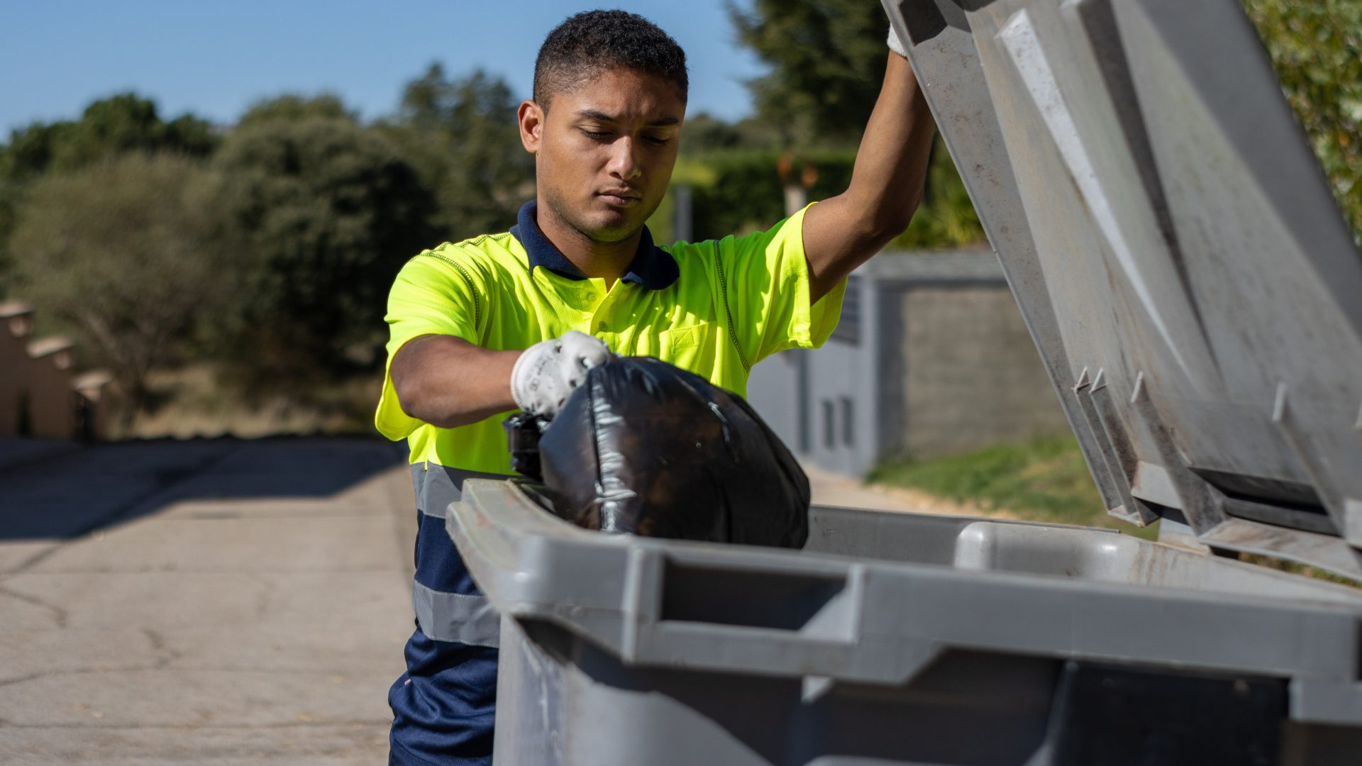 A person in a neon green shirt puts a black trash bag in a gray bin outdoors.