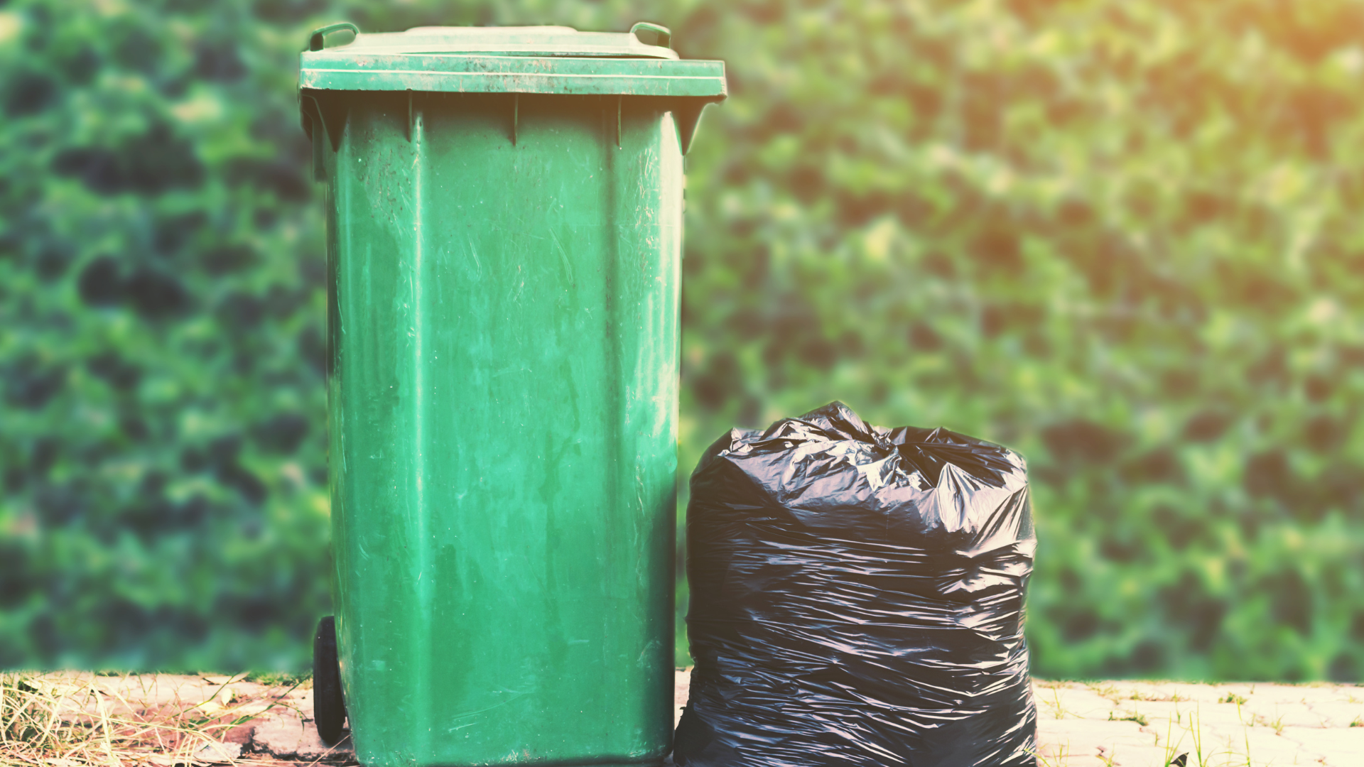 A green trash can and a black trash bag are sitting next to each other.