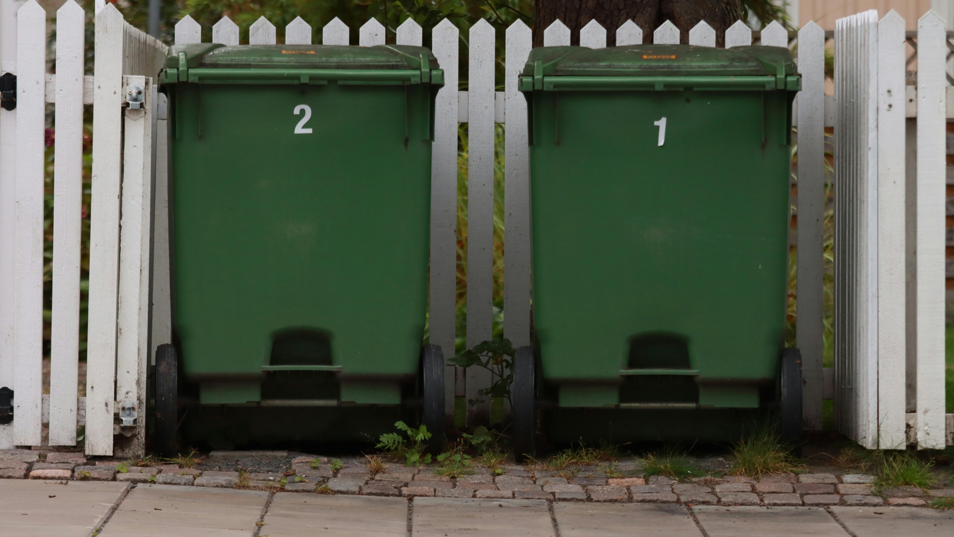 Two green trash bins numbered 1 and 2 next to a white picket fence.