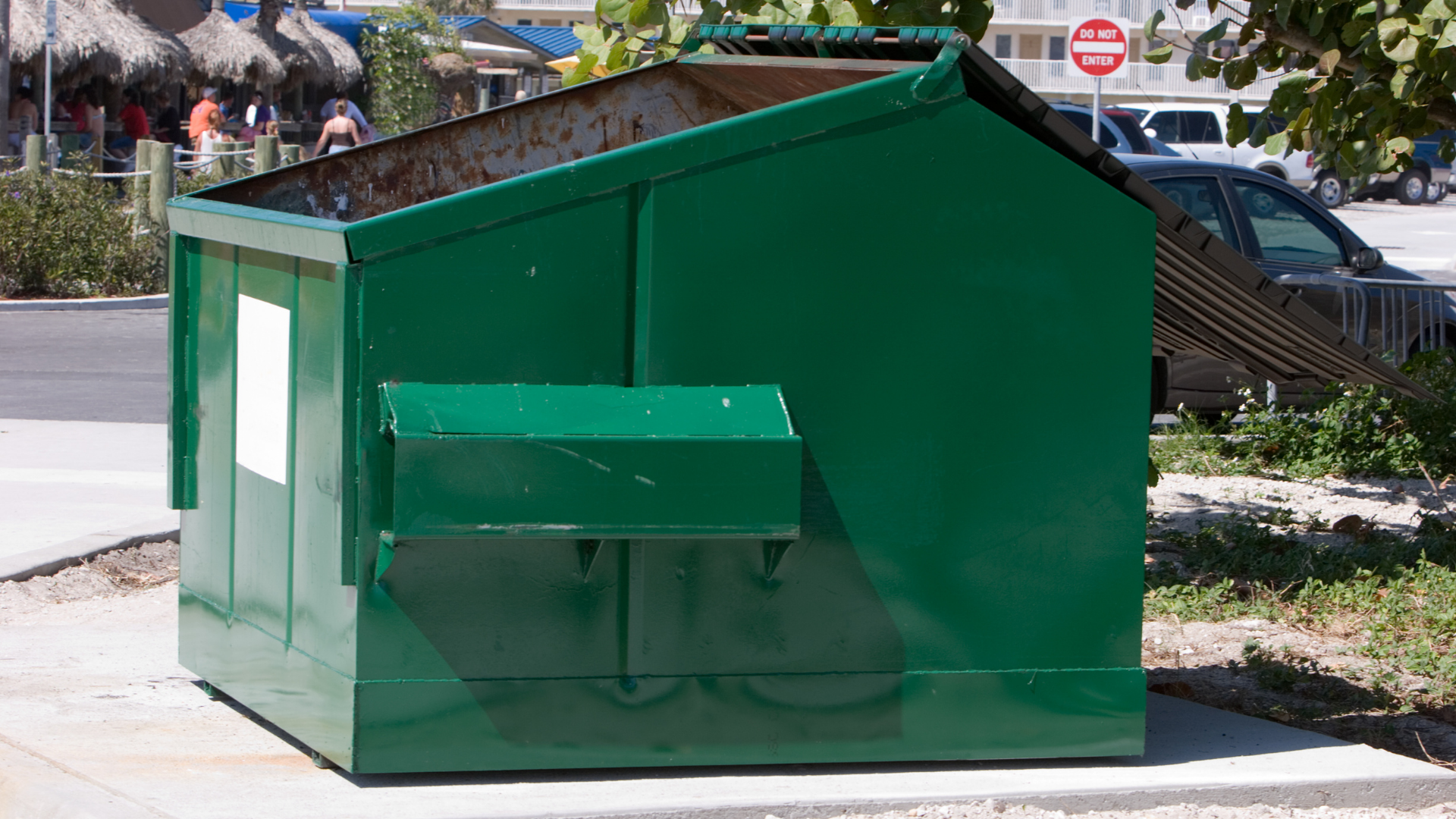Green dumpster on a sidewalk, near a street with parked cars and a 
