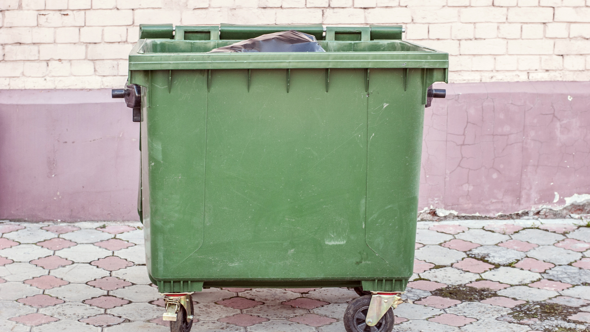 Green trash dumpster on wheels, filled with waste, set against a brick wall and patterned ground.