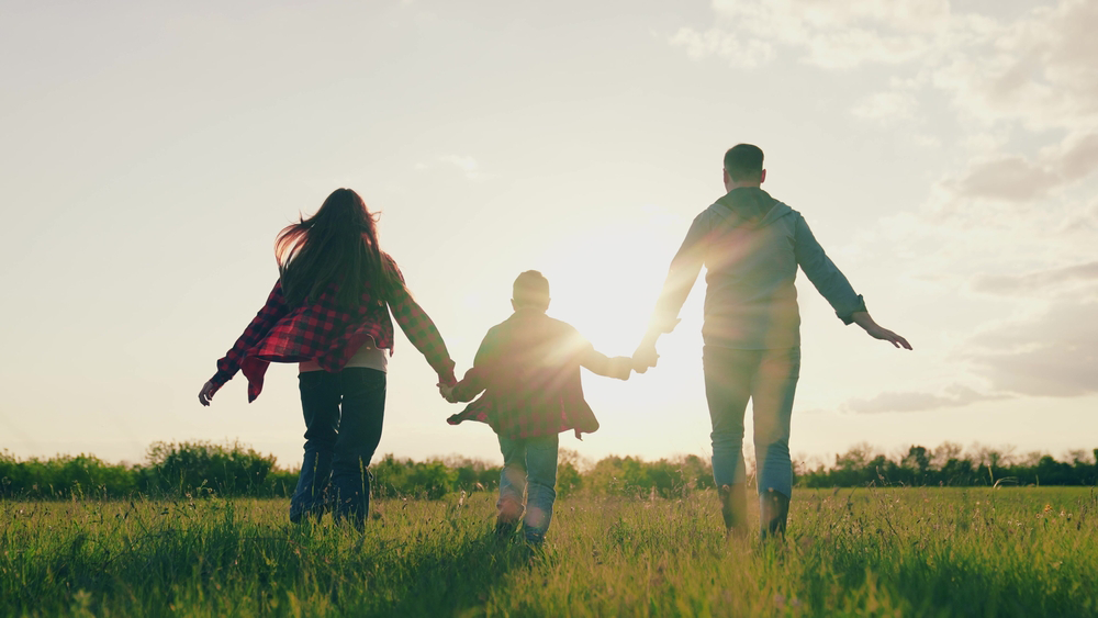 Family holding hands, running across a grassy field towards the sun.
