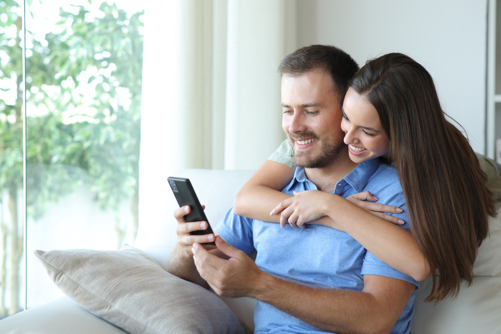 Couple looking at phone together, smiling on a couch near a window.