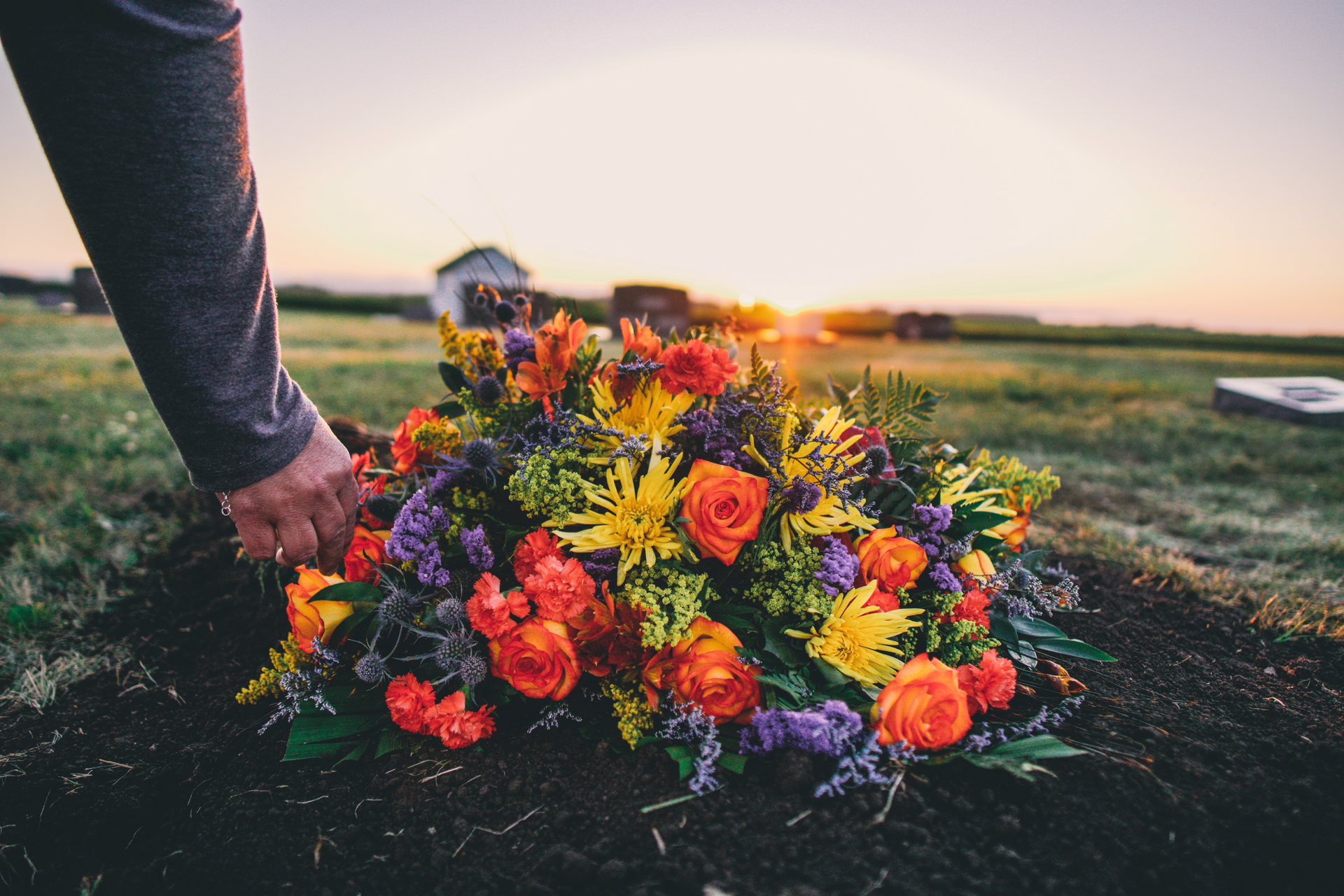 Person placing colorful floral arrangement on a grave in a cemetery at sunset.