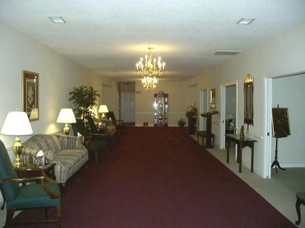 Long hallway with red carpet, ornate chandelier, couches, and doorways, likely a funeral home.