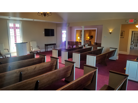 A chapel with wooden pews, red carpet, and a podium. Beige walls, windows, and a doorway are visible.