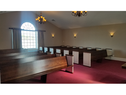 Interior of a chapel with wooden pews, red carpet, and a large arched window with natural light.