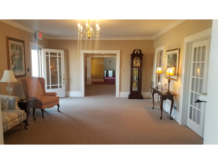 A spacious room with beige walls and carpet. A grandfather clock stands next to a table with lamps. Doors on either side.