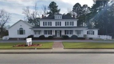 White two-story building with a porch, surrounded by greenery and trees. A sign is in front.