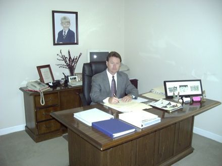 Man in a suit sits at a desk in an office. A photo hangs on the wall, and the desk has papers.