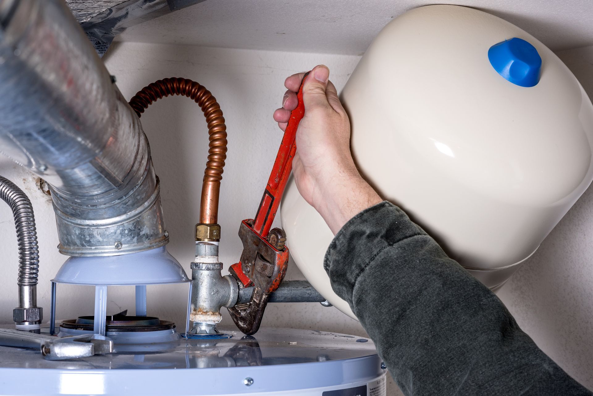 A hand tightens a water heater pipe with a red wrench, the water heater being next to a wall.