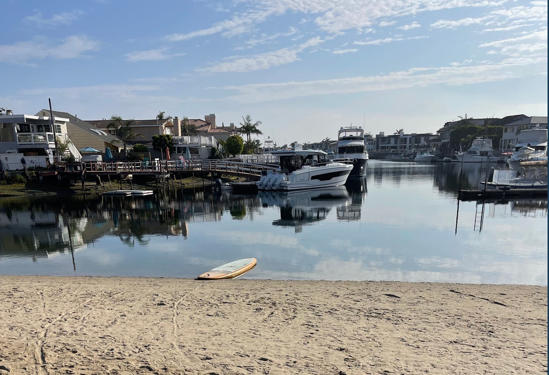 Sandy beach overlooking calm water with boats docked in harbor under a blue sky.