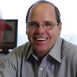 A man wearing glasses and a grey shirt smiles for the camera