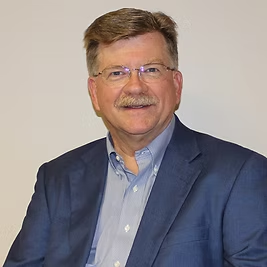 A man in a suit and glasses is sitting in front of a white wall.