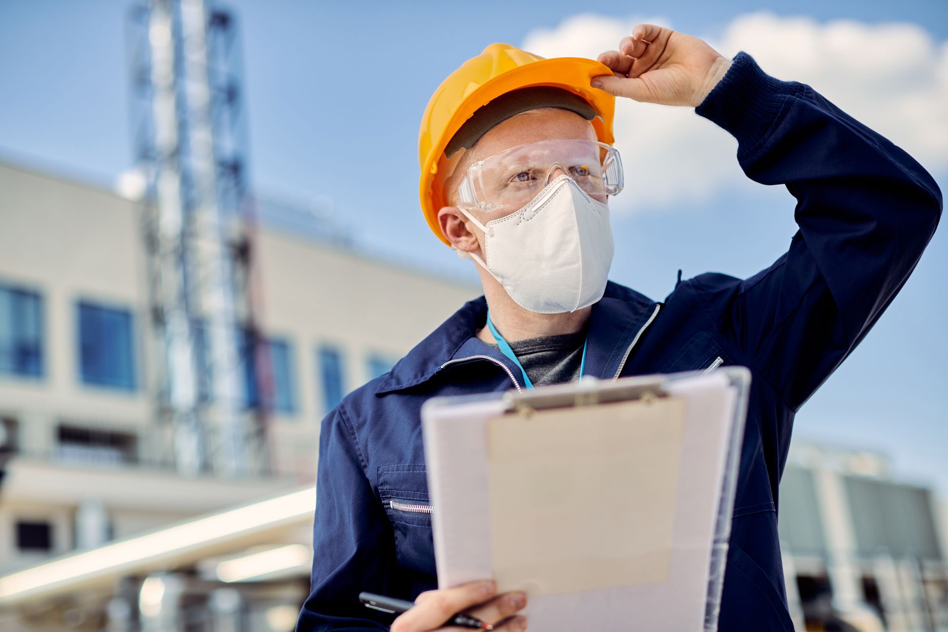 A man wearing a hard hat and a mask is holding a clipboard. 