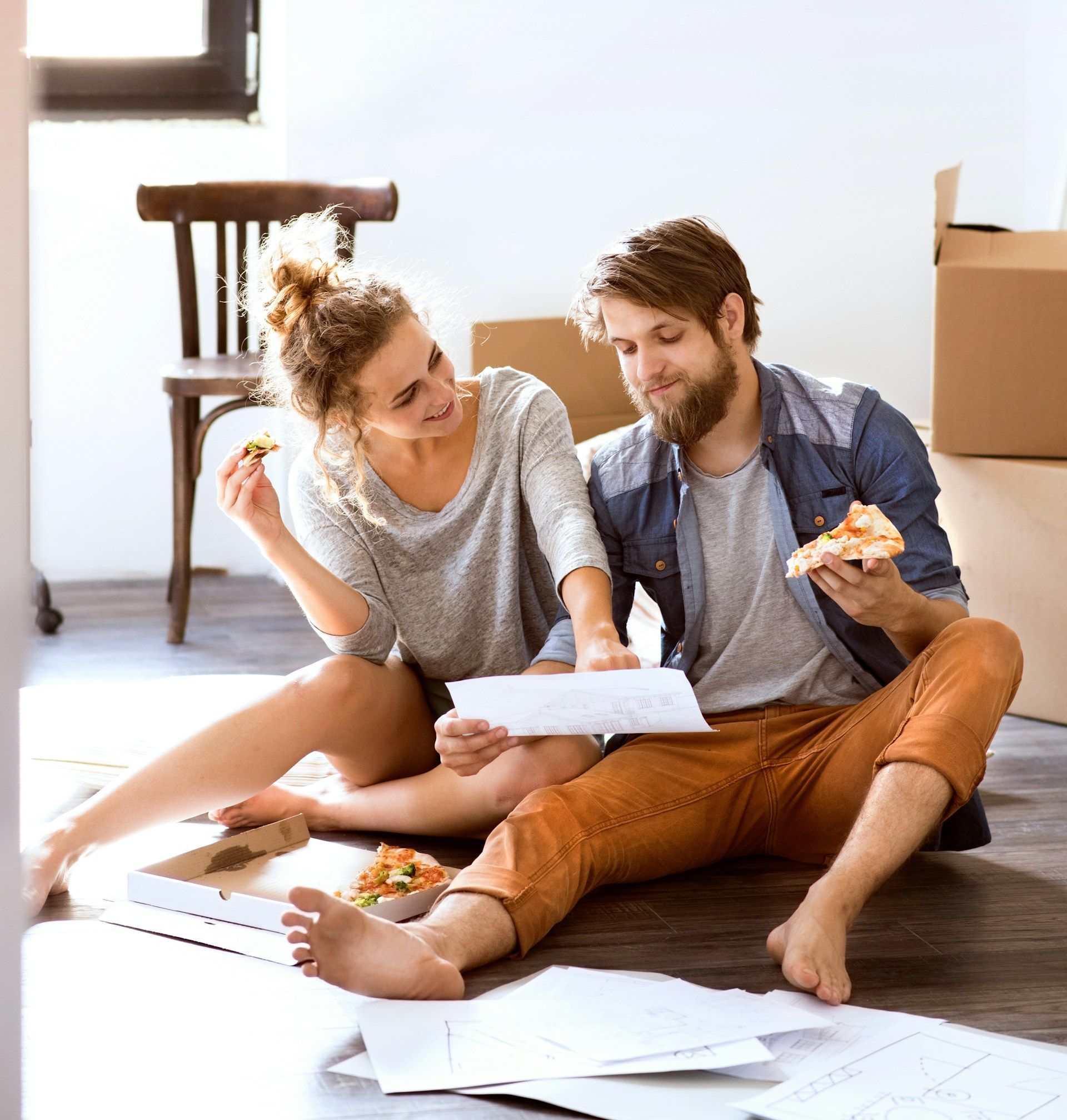 A man and a woman are sitting on the floor eating pizza. 