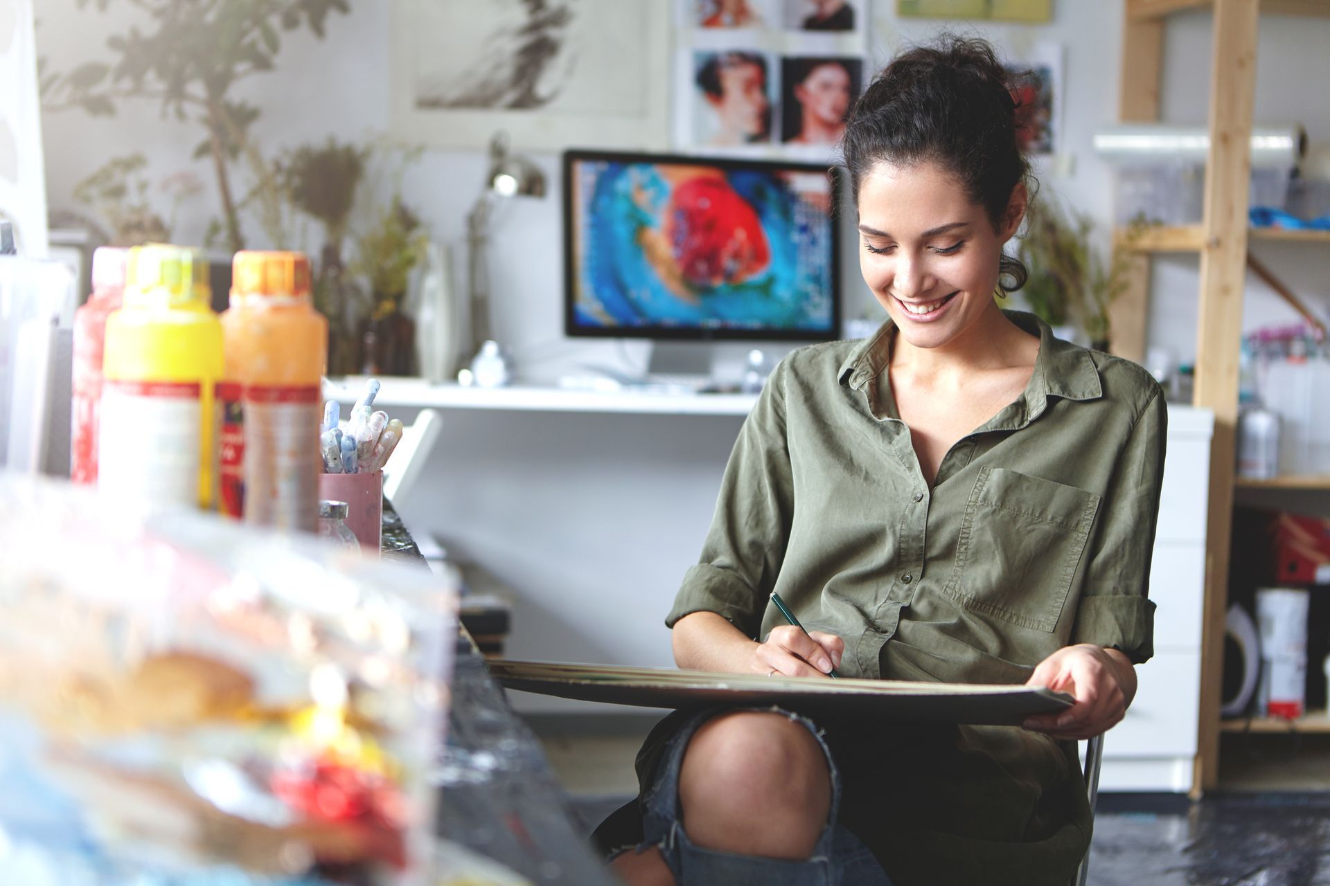 A woman is sitting on the floor in an art studio looking at a painting. 