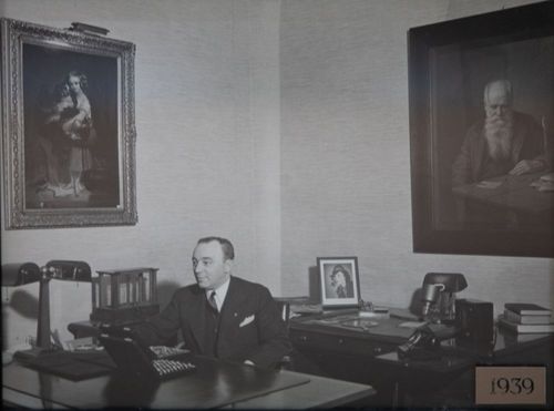 Man seated at a desk in an office with framed portraits on the wall, dated 1939.