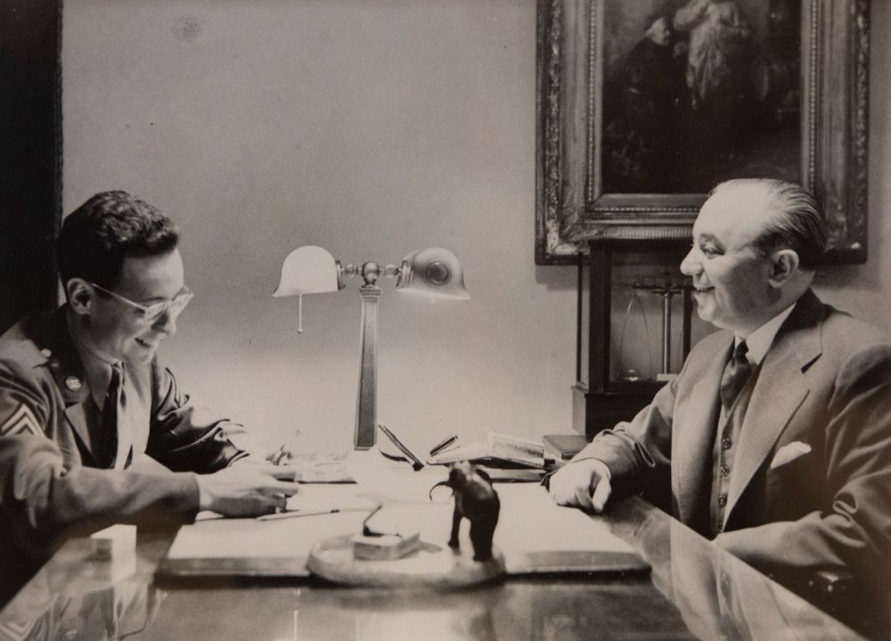 Two men in suits sit across a desk in a formal office, talking under a desk lamp.