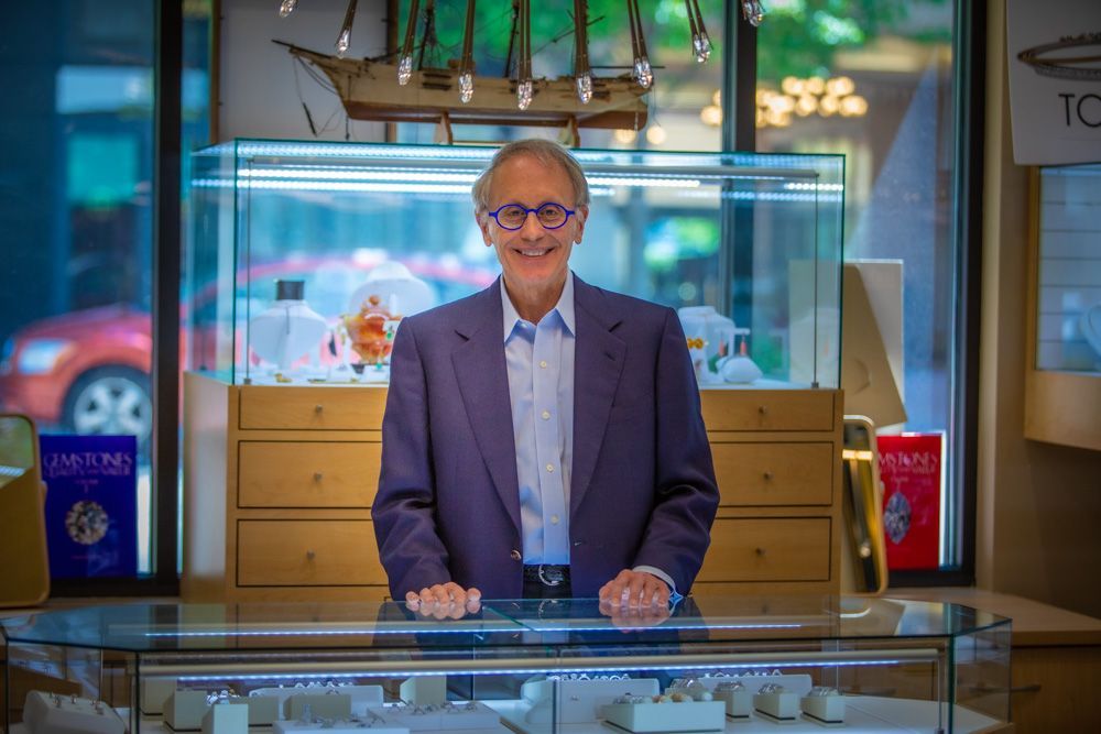 Man standing behind a glass display counter in a shop, smiling in a purple blazer and blue shirt.