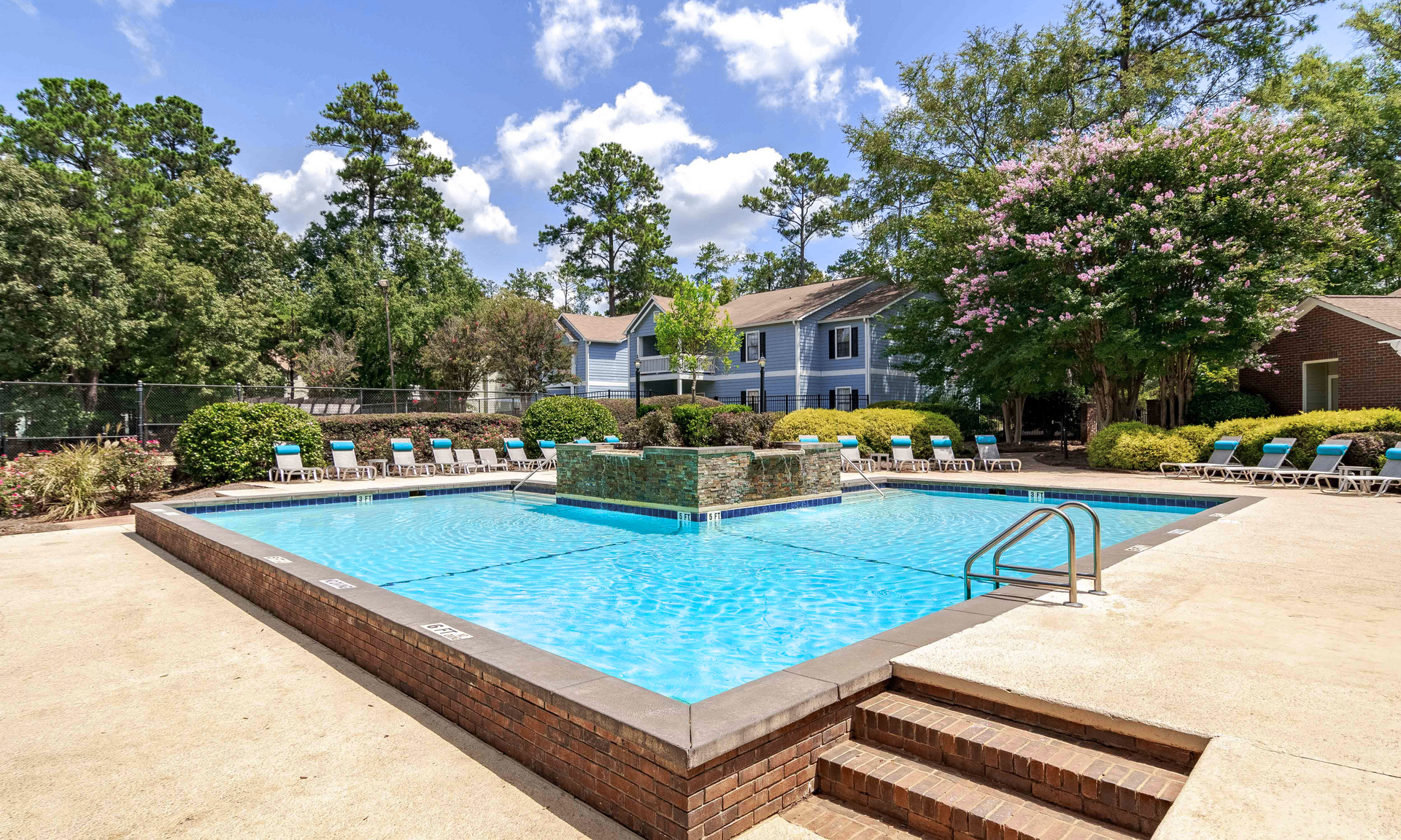 Swimming pool at an apartment complex. Blue water, brick border, lounge chairs, trees and blue-gray buildings in the background.