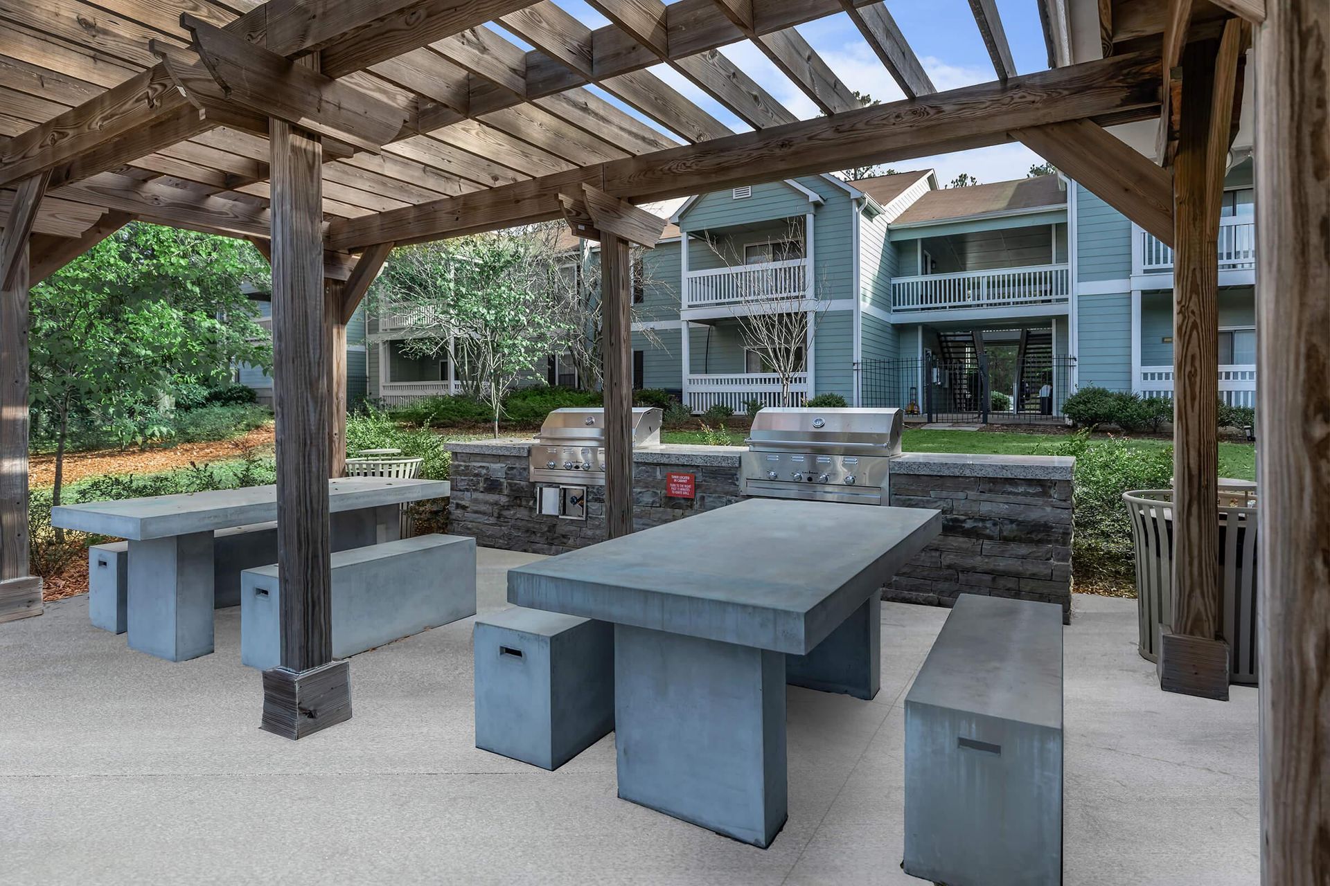 Outdoor grilling area with tables and benches under a wooden pergola, with apartment buildings in the background.