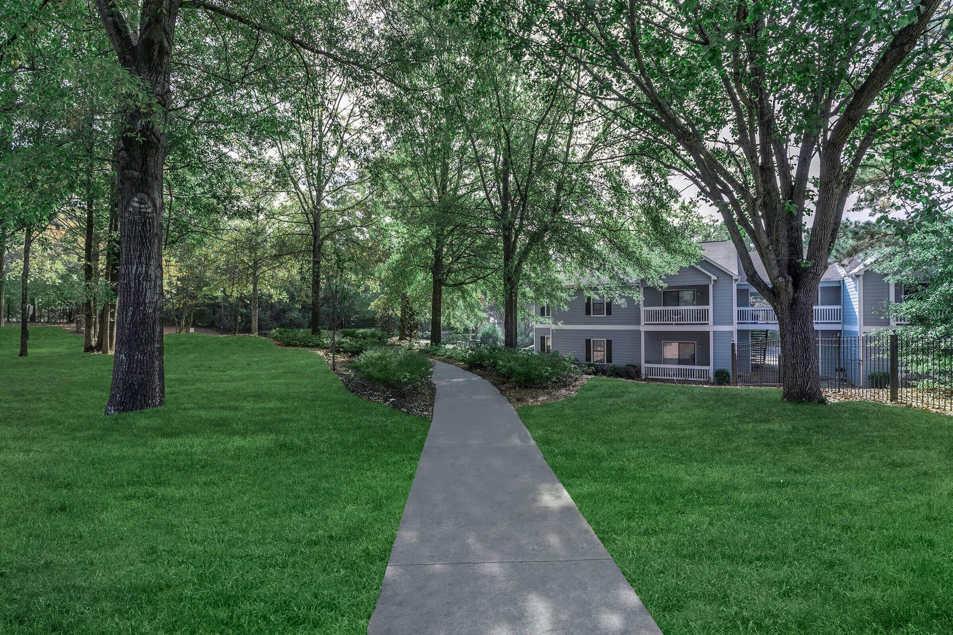 A paved path through a grassy yard leads to a two-story apartment building surrounded by trees.