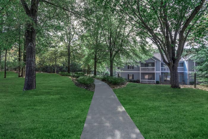 A paved path through a grassy yard leads to a two-story apartment building surrounded by trees.