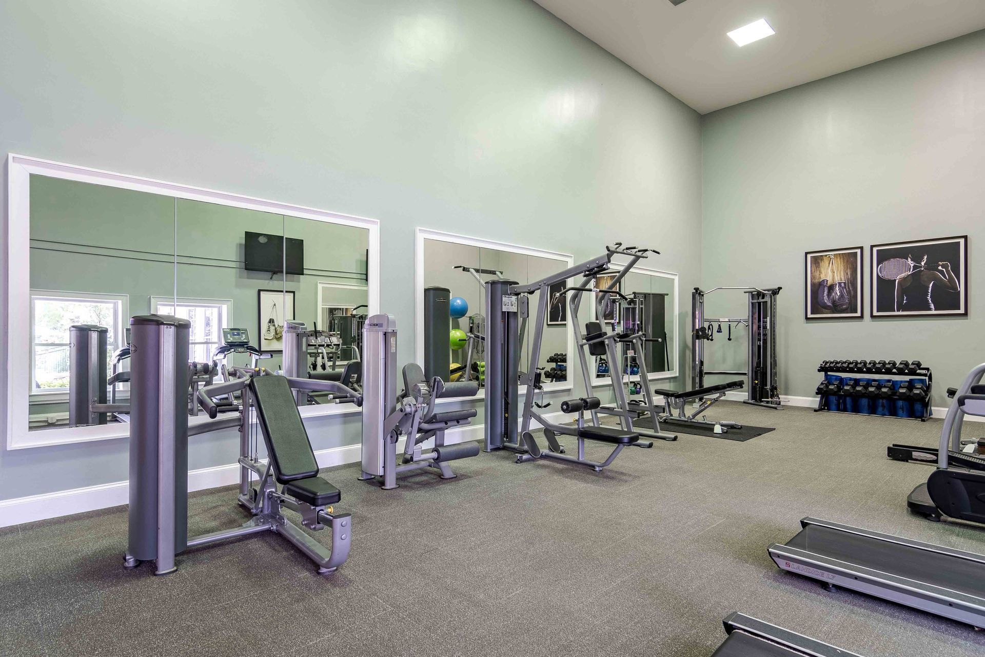 Gym with weight machines, benches, and free weights on gray floor. Large mirrors, light walls.