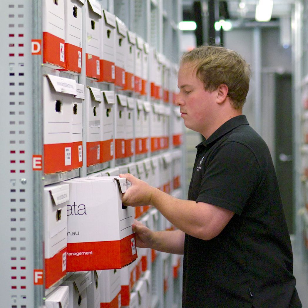 Man in black shirt retrieving a box from a shelf filled with labeled storage boxes. White and red boxes in a file room.