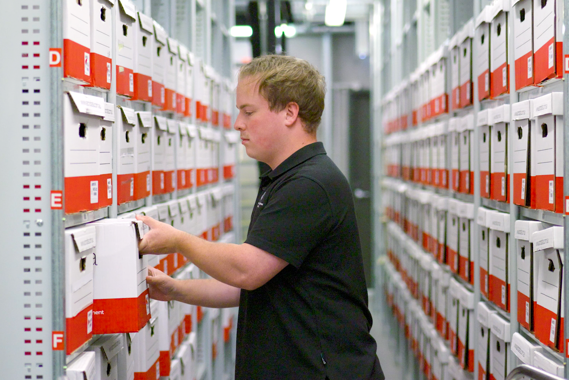 Man in black shirt shelving a file box in a storage room with rows of boxes on shelves.