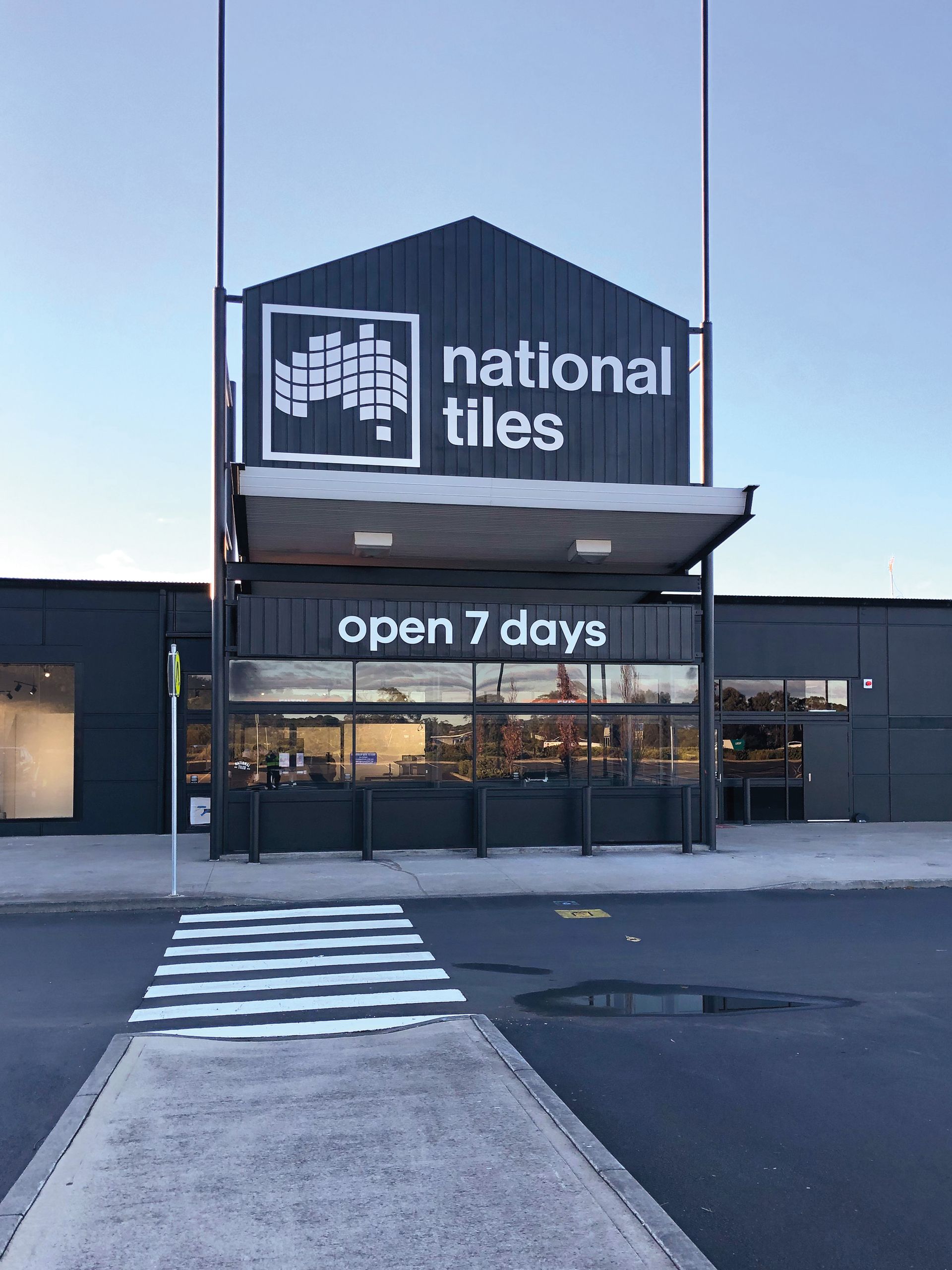 Exterior of a National Tiles store with the sign “open 7 days”. A crosswalk is in the foreground.