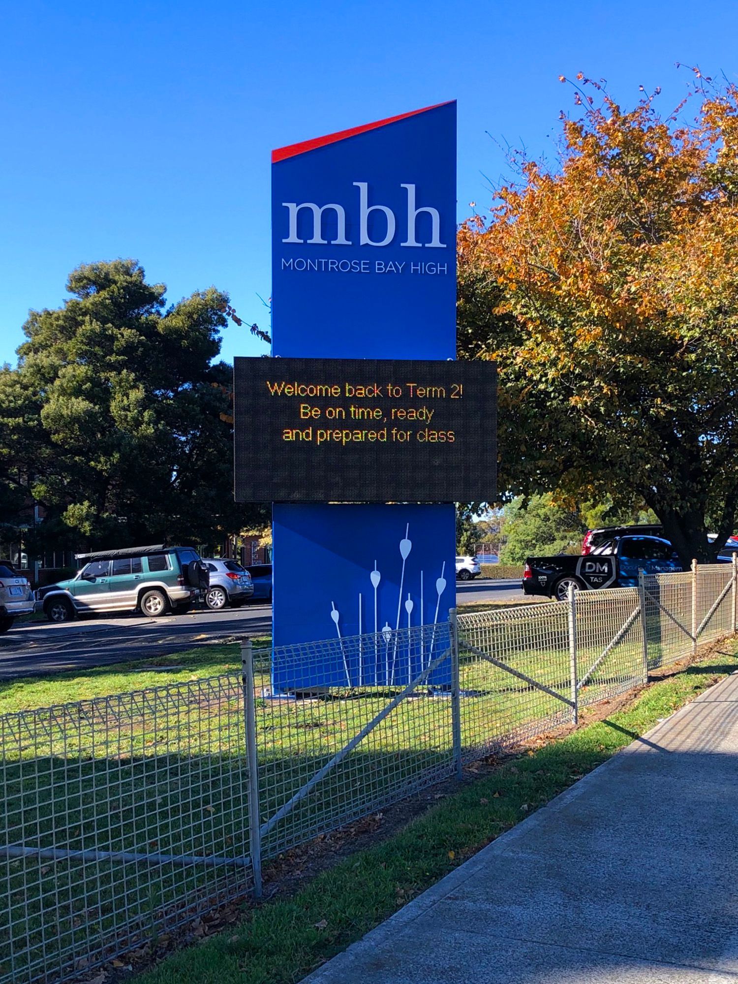 Sign for MBH (presumably a school), blue with white text and logo; shows a welcome message and silhouettes of plants.