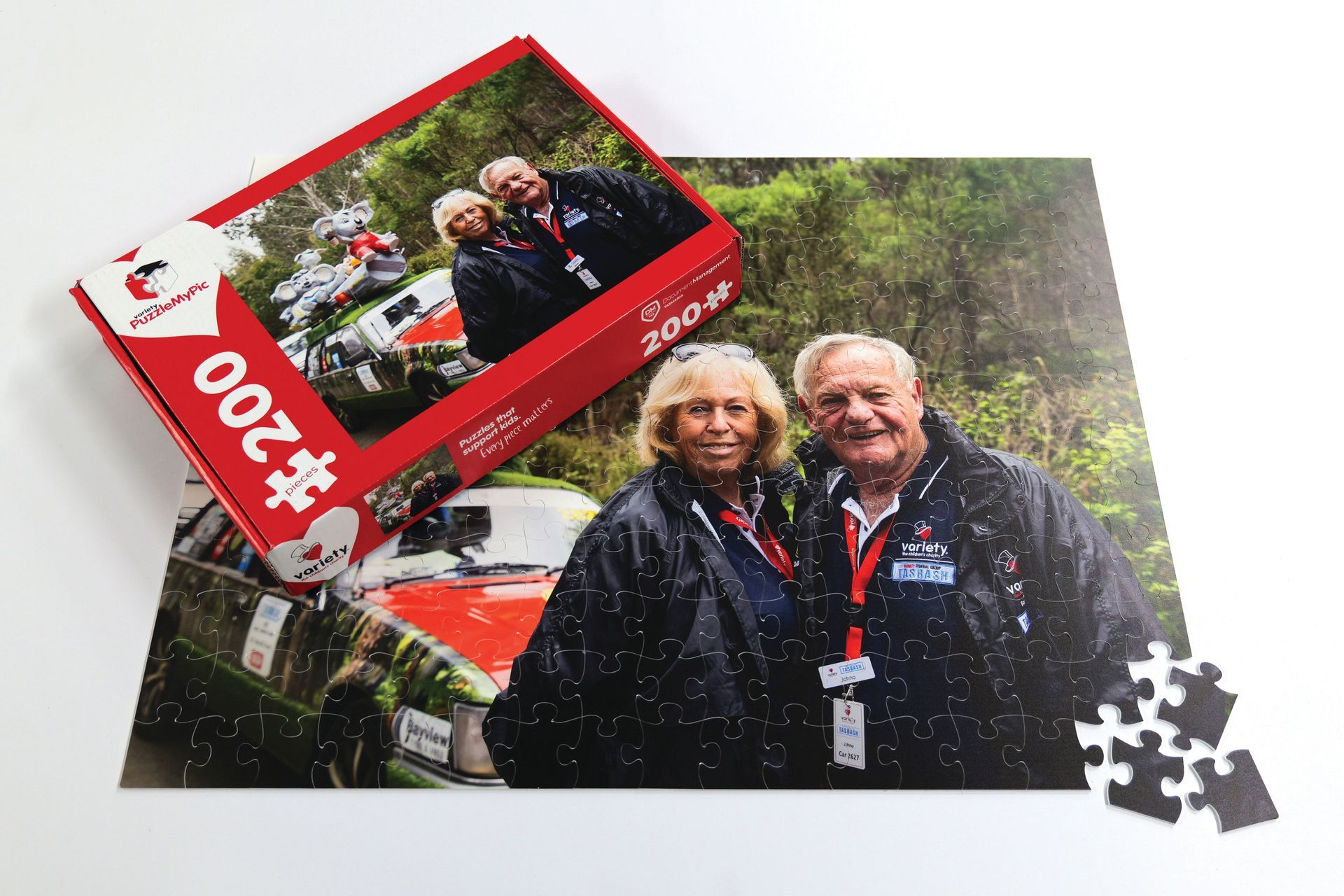 A couple smiles, standing in front of a red race car. A puzzle box and scattered pieces are present.