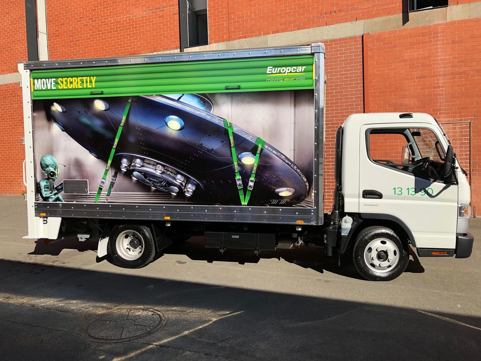 White truck with an image of a UFO secured inside. Green straps and background. Brick building.