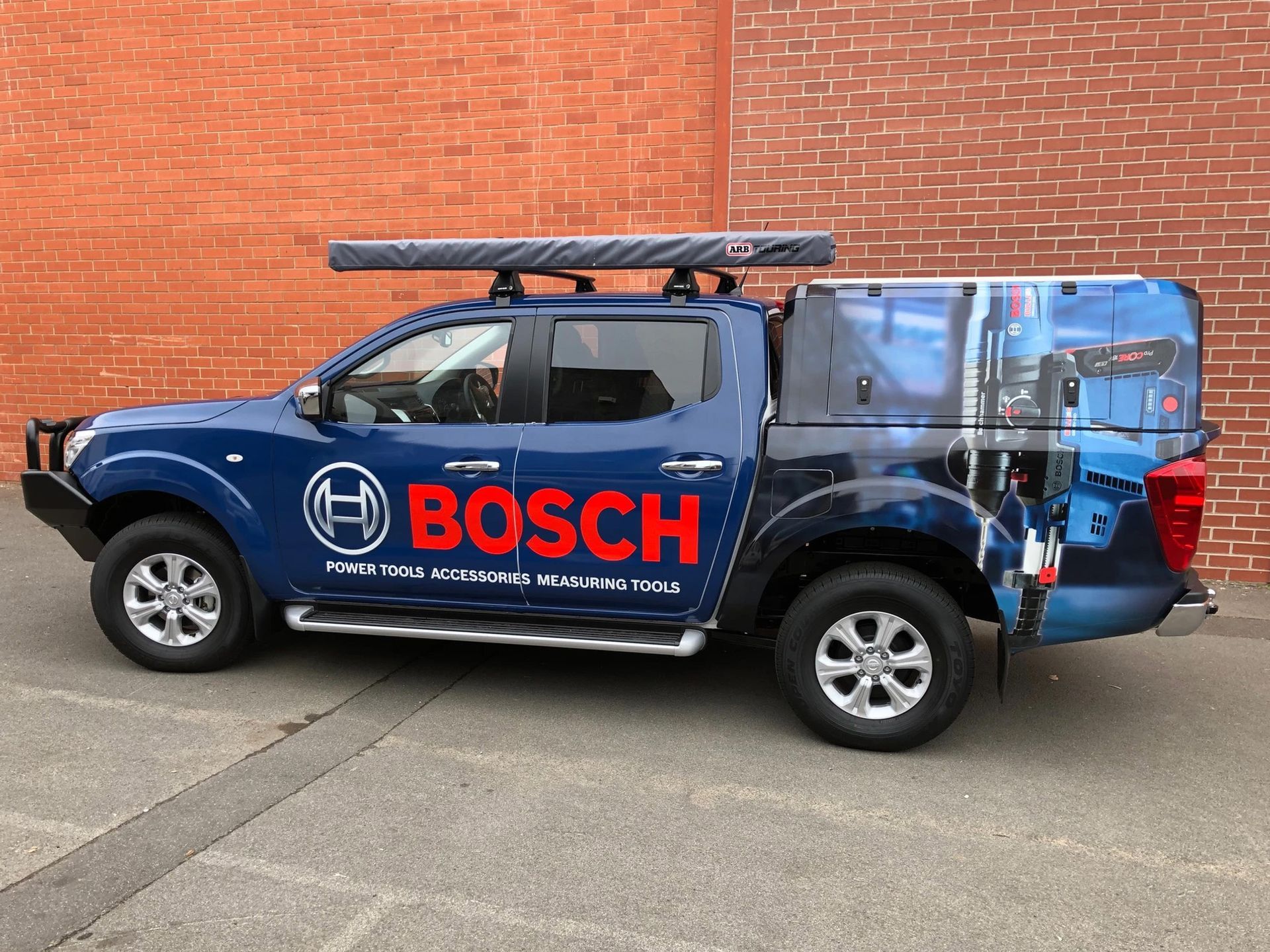 Blue Bosch branded pickup truck with a roof rack and camper shell parked in front of a brick wall.