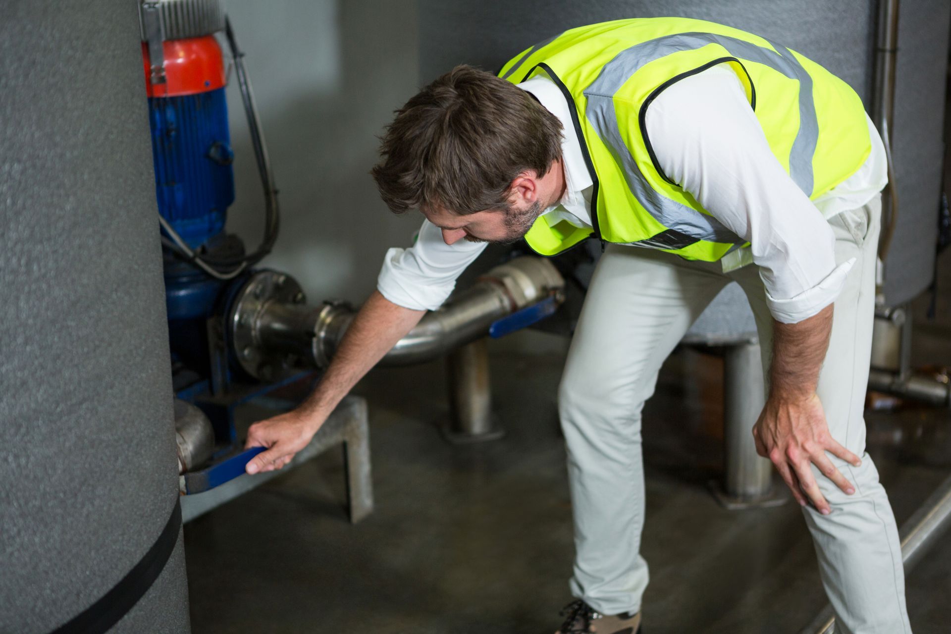 A man in a yellow vest is kneeling down and looking at a pipe.