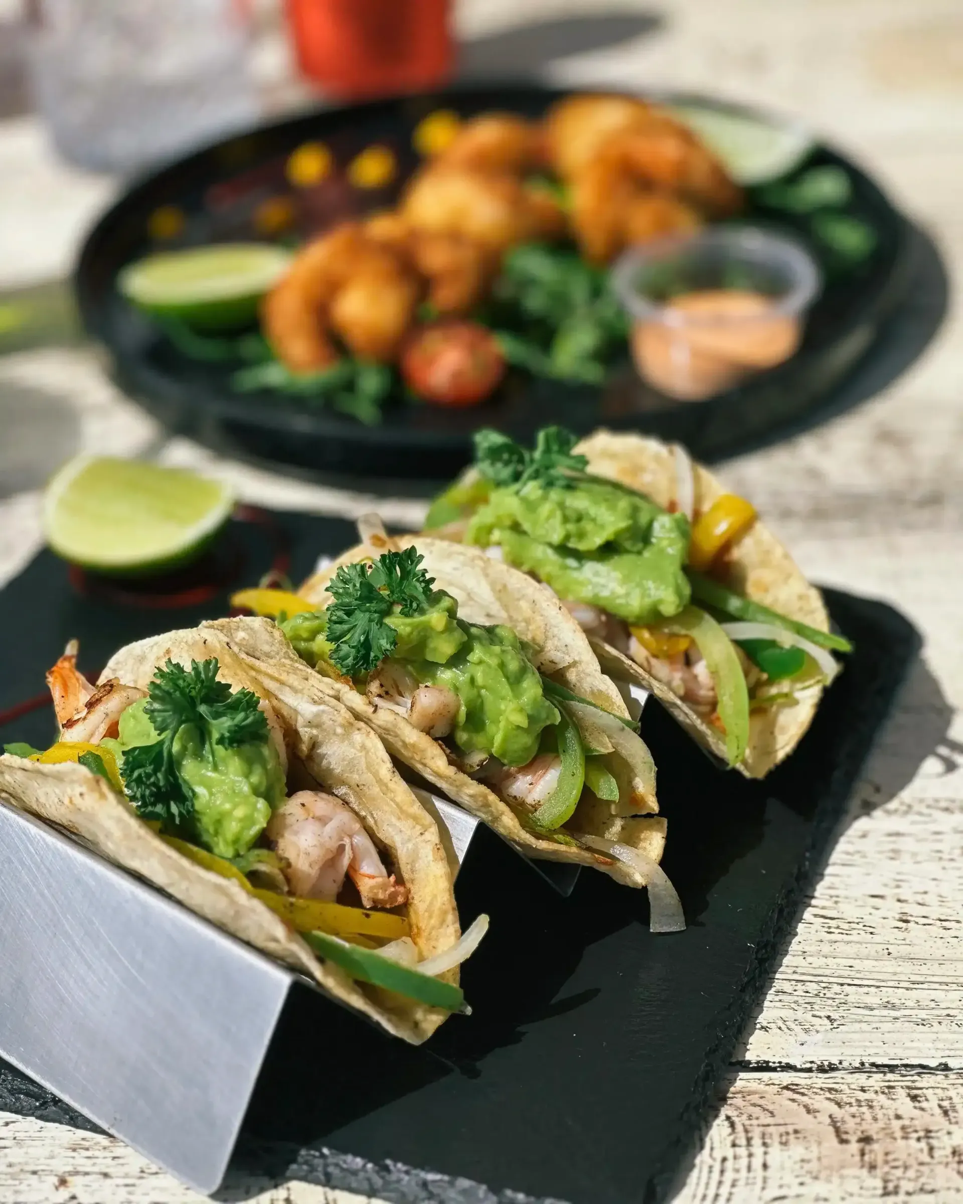 Hay tres tacos en una bandeja de pizarra sobre una mesa al lado de un plato de camarones.