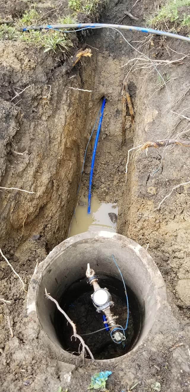 An open well with equipment inside and blue wires coming out of the hole, set in the dirt.