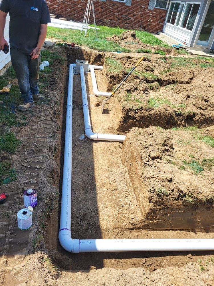 A person stands near a trench holding PVC pipes, connecting them in a yard near a house.