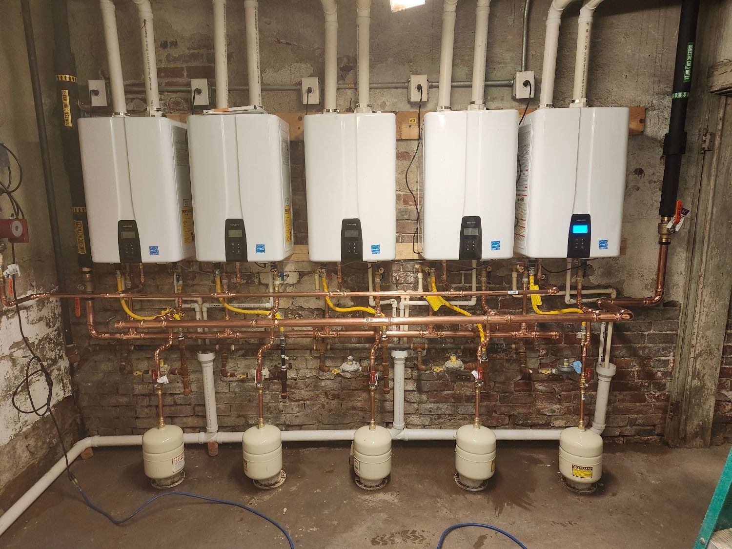 Five white tankless water heaters mounted on a brick wall with copper pipes and white drainage.