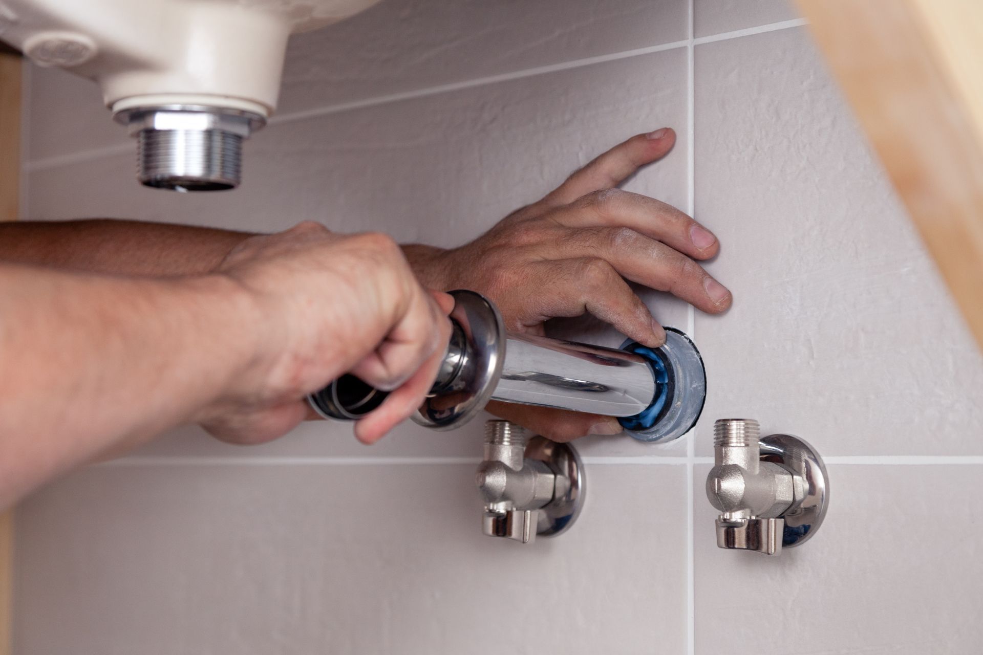 Hands installing chrome plumbing under a sink in a bathroom with beige tiles.