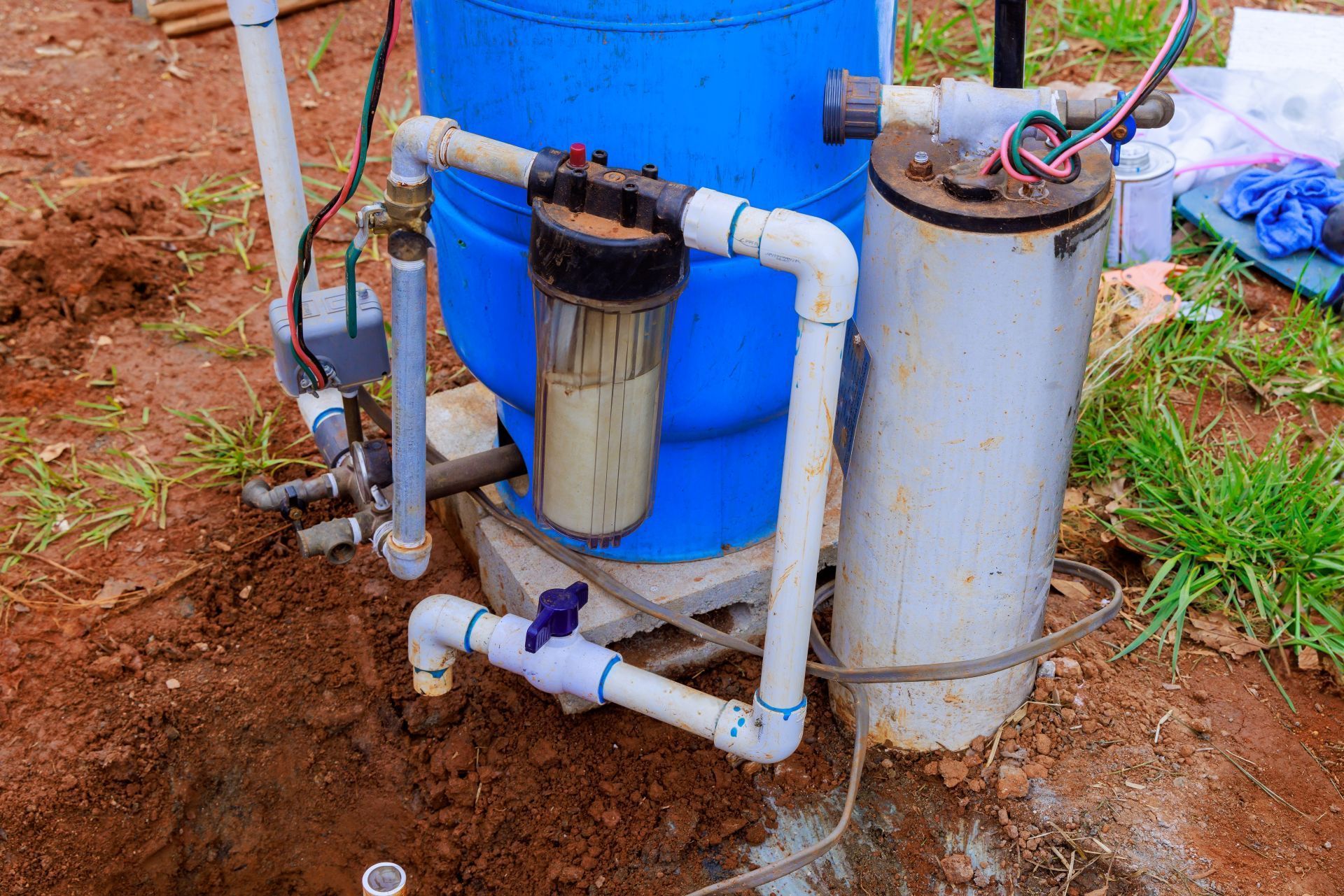 Blue water tank and plumbing components outdoors in a partially dug hole.